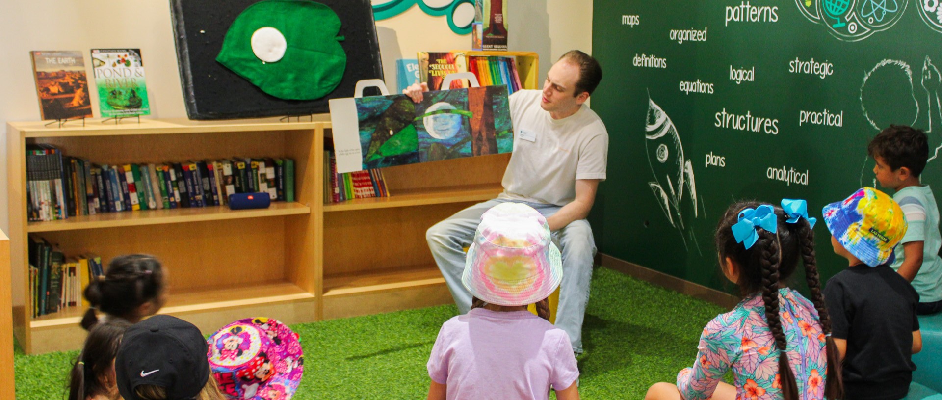 Guy is holding up a book while reading it to children sitting around him.