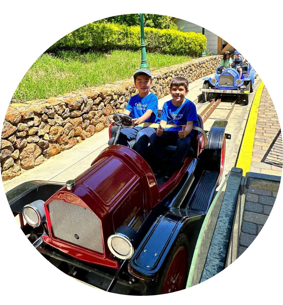 Two boys in blue shirts smiling while driving a red car on a sunny day.